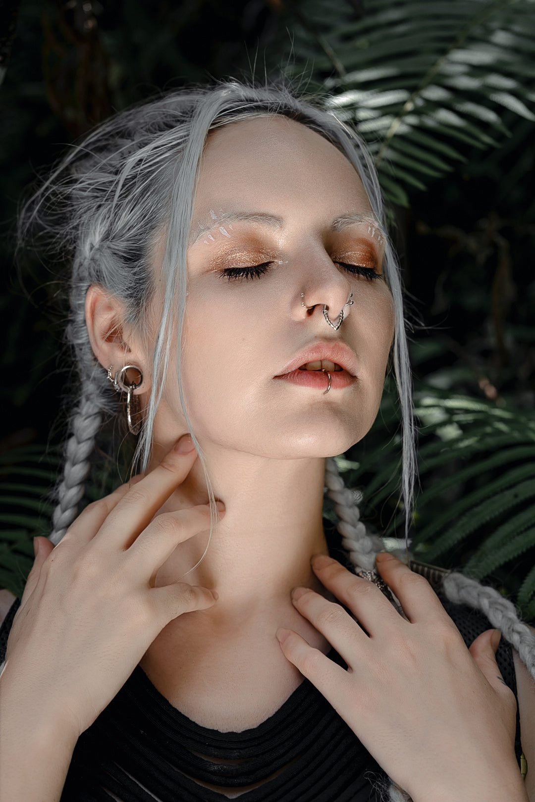 A person with light gray braided hair and several handcrafted body jewelry, including a Kaya Movement's KAMO Biomech Teardrop Clicker Septum Ring in Silver (16 gauge) and a lip ring, closes their eyes while touching their neck with both hands. They are surrounded by green foliage, creating a serene, natural background.